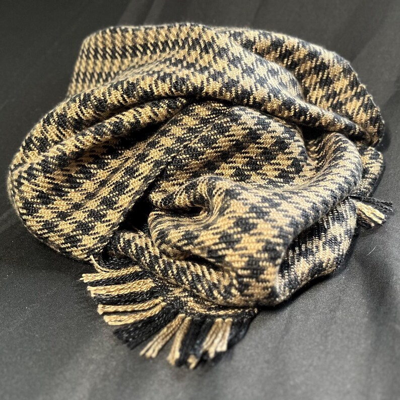 May include: A folded scarf with a black and tan houndstooth pattern. The scarf has a fringed edge in matching colors. The fabric appears to be soft and warm, suitable for cold weather.