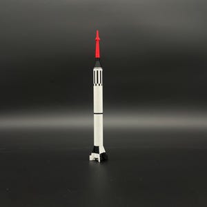 May include: A white and black model rocket with a red nose cone. The rocket has black stripes and a black base. The rocket is standing upright against a dark background.