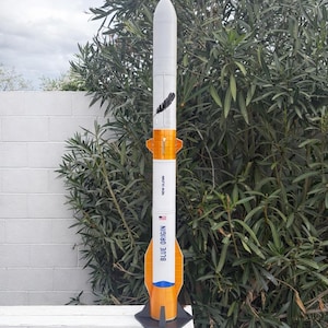 May include: A model rocket with a white and orange design, featuring the text "BLUE ORIGIN" and "NEW GLENN". A black feather graphic is on the side. The rocket is mounted on a black stand, set against a backdrop of greenery and a white fence.