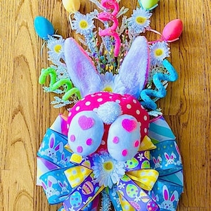 May include: A pink and white bunny-shaped Easter wreath with blue, yellow, and purple ribbon. The wreath is decorated with pastel-colored eggs and white flowers.