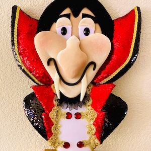 May include: A decorative wall hanging in the shape of a cartoon vampire. The vampire has a black cape and hair, a pale face, and red sequined accents. The vampire's vest has gold trim and red jewels.
