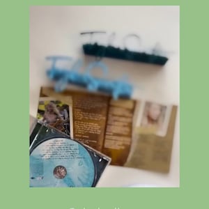 May include: A blue CD with a white and blue design. The CD is in a clear plastic case. The case is open and the CD is visible. The CD is on a table with a book and a blue hanger.