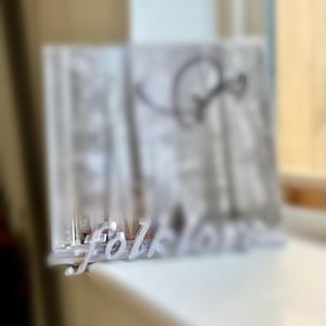May include: A clear acrylic display stand with the word "Folklore" in a cursive font. The stand holds a blurred image of a bicycle and trees. The stand is on a white surface.