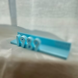 May include: A light blue, three-dimensional sculpture of the numbers "1989" in a stylized font. The numbers are attached to a rectangular base with a groove. The background is blurred.
