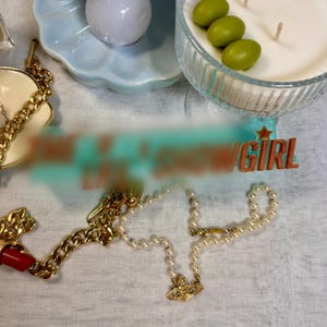 May include: A gold chain necklace with a red lipstick charm and a pearl necklace with a gold pendant. The image also includes a gold chain bracelet, a candle, and a decorative plate. The word "GIRL" is visible.