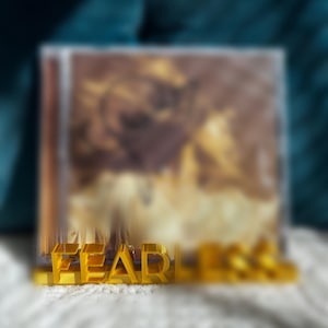 May include: A gold-colored, three-dimensional word "FEAR" is displayed in front of a blurred background. The background appears to be a framed image with brown and yellow tones. The word "FEAR" is the focal point.