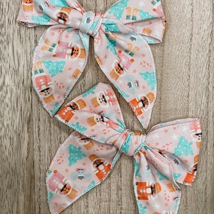 May include: Two pink fabric hair bows with a Christmas nutcracker pattern. The bows are tied together and have a white trim.