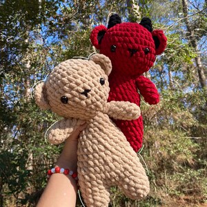 May include: Two crocheted plush toys, one red with horns and one beige, are held in a hand. The red toy has black eyes and a black nose. The beige toy has black eyes and a black nose. Both toys have a stitched smile.