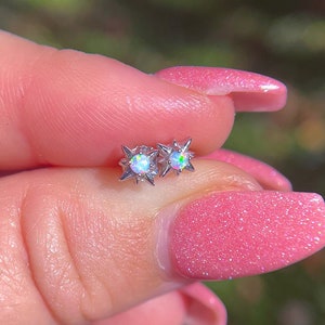 May include: A pair of silver star-shaped earrings with opal stones. The earrings are held in a hand with pink nail polish.