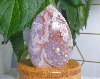 Puede incluir: Una piedra de ágata rosa y blanca con un patrón único, descansando sobre un soporte de bambú natural. La piedra tiene una superficie lisa y pulida y es un hermoso ejemplo de belleza natural.