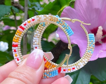 May include: A pair of colorful beaded hoop earrings with a gold tone finish. The earrings are made with small, colorful beads in a geometric pattern. The earrings are shown against a background of green leaves and pink flowers.