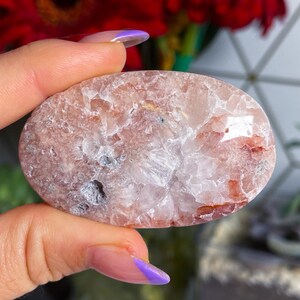 Puede incluir: Un cristal de ágata liso, de forma ovalada, rosa y blanco con una textura natural y rugosa. El cristal tiene una apariencia translúcida con un toque de rojo.
