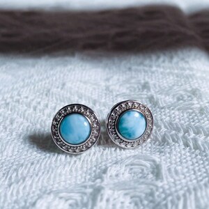 May include: A pair of silver earrings with a round blue gemstone set in a silver bezel. The earrings are on a white fabric background.