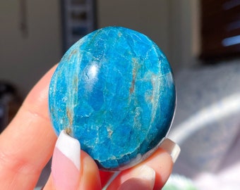 May include: A smooth, polished blue apatite crystal sphere. The crystal has a light blue color with darker blue streaks throughout. The sphere is being held in a hand.