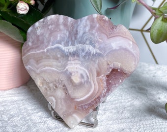 May include: A heart-shaped, polished agate stone in shades of pink, white, and lavender, displayed on a silver stand. The stone has a geode-like interior with crystal formations. The background includes potted plants with green and purple foliage.