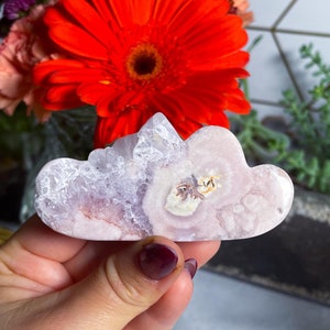 Puede incluir: Un cristal de amatista de color rosa pálido y blanco tallado en forma de nube. El cristal tiene una textura natural, rugosa y es translúcido.