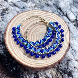 May include: A pair of gold hoop earrings with blue and light blue beaded fringe. The earrings are on a wooden surface.