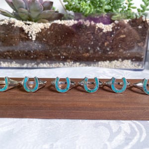May include: Six silver rings with turquoise horseshoe designs. The rings are arranged in a row on a wooden surface.