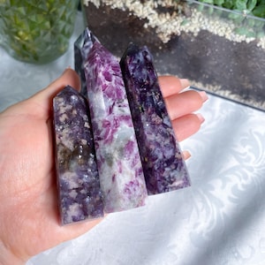 May include: Four purple and white crystal wands, also known as points, are held in a hand. The wands are approximately 5 inches long and have a pointed tip.