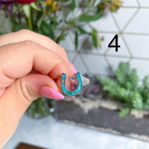 May include: A silver ring with a turquoise horseshoe design. The ring is on a finger.