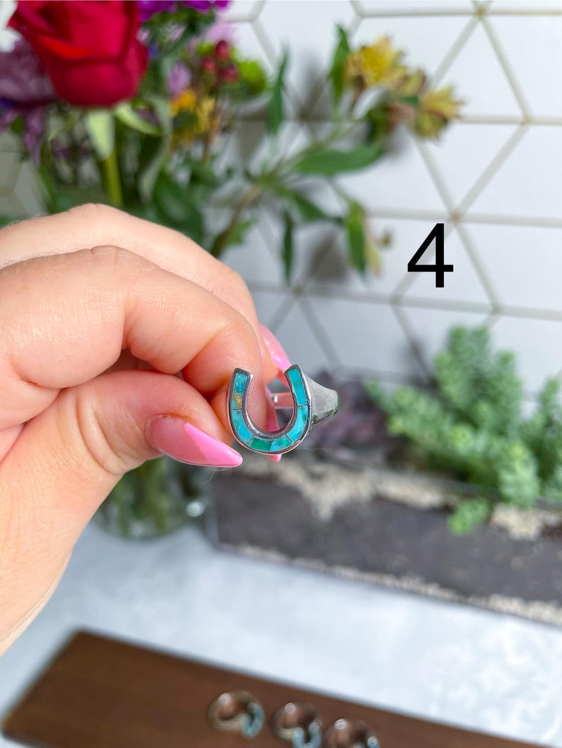 May include: A silver ring with a turquoise horseshoe design. The ring is on a finger.