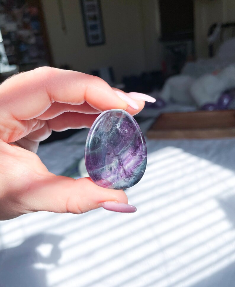 May include: A smooth, oval-shaped fluorite crystal with purple, green, and white swirls. The crystal is polished and has a translucent appearance.