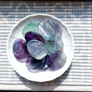 May include: A collection of polished fluorite stones in various shades of purple, green, and blue, arranged on a white plate.
