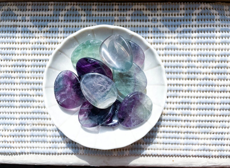 May include: A collection of polished fluorite stones in various shades of purple, green, and blue, arranged on a white plate.