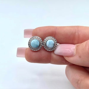 May include: A pair of silver earrings featuring a round, light blue gemstone set in a silver bezel. The earrings have a decorative border around the gemstone.