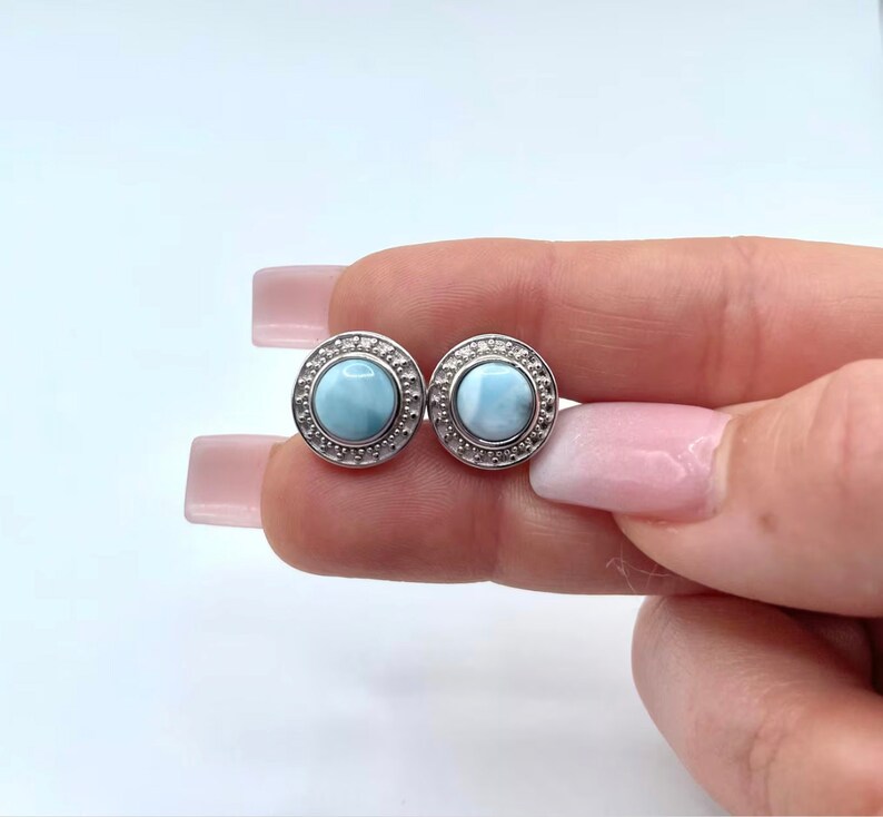 May include: A pair of silver earrings featuring a round, light blue gemstone set in a silver bezel. The earrings have a decorative border around the gemstone.