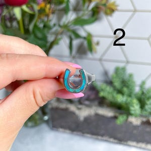 May include: A silver ring with a turquoise horseshoe design. The ring is on a finger and the number 2 is visible in the background.