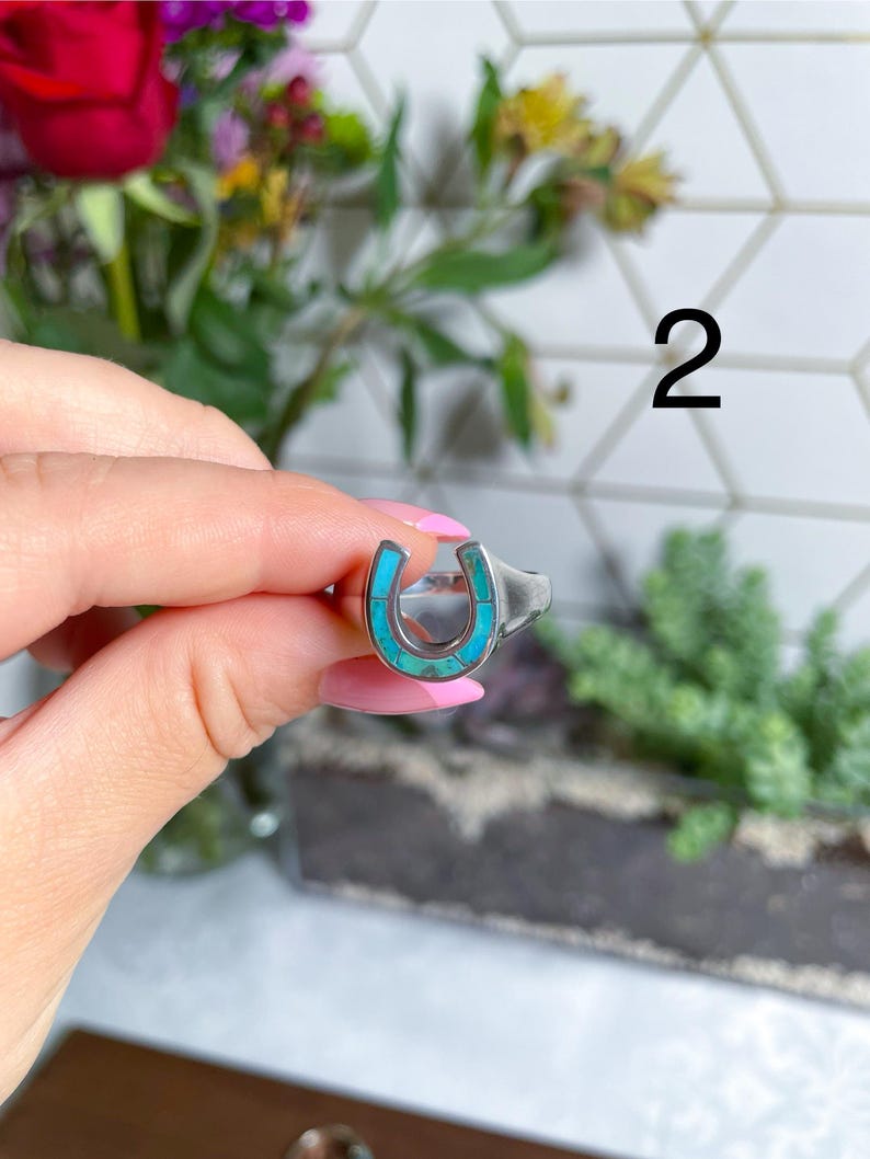 May include: A silver ring with a turquoise horseshoe design. The ring is on a finger and the number 2 is visible in the background.