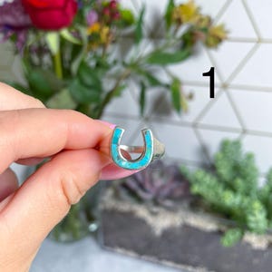 May include: A silver ring with a turquoise horseshoe design. The ring is on a finger.