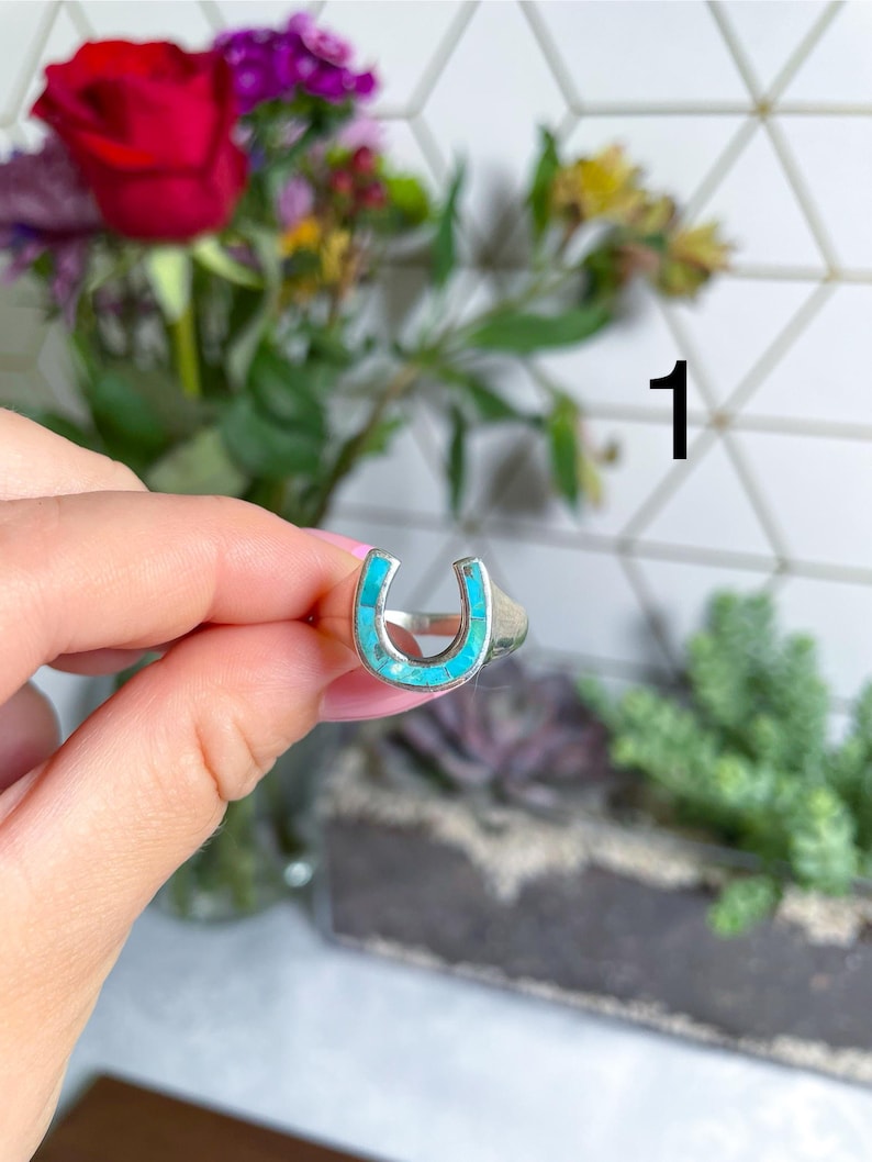 May include: A silver ring with a turquoise horseshoe design. The ring is on a finger.