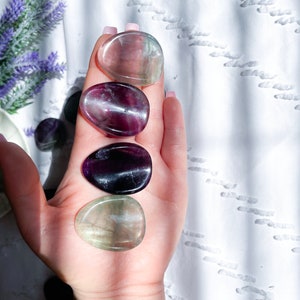 May include: Five smooth, polished fluorite stones in various shades of green, purple, and black. The stones are arranged in a row on a hand, with the green stones at the top and bottom.