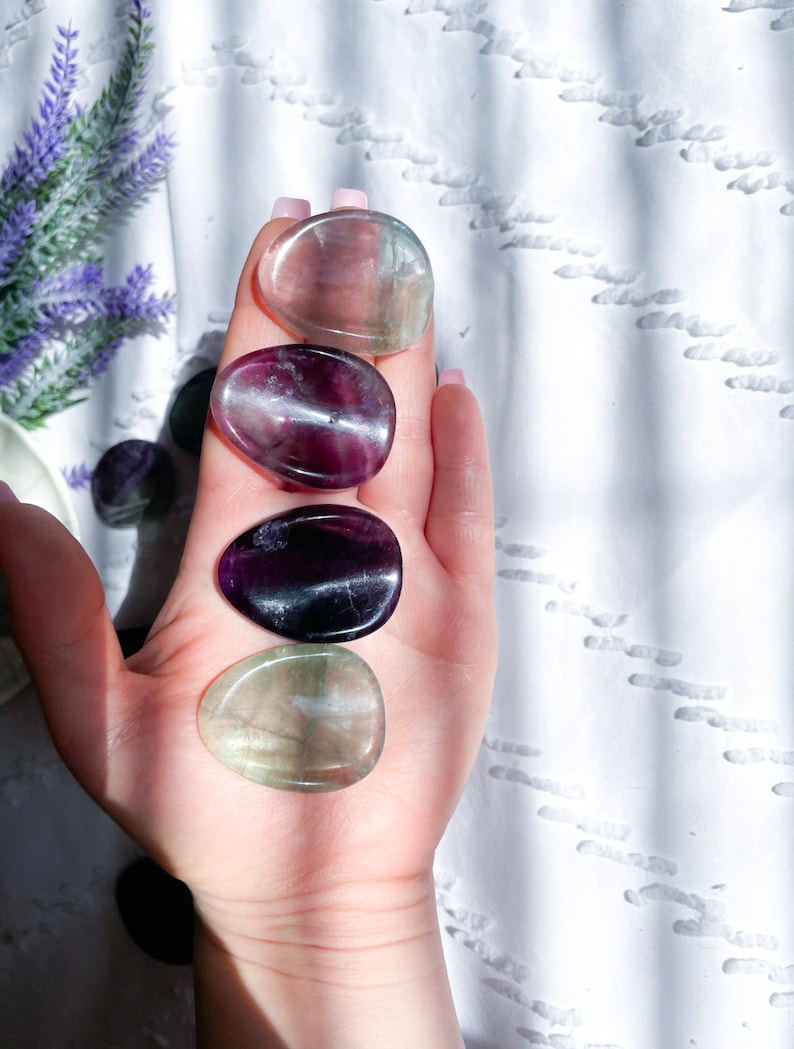 May include: Five smooth, polished fluorite stones in various shades of green, purple, and black. The stones are arranged in a row on a hand, with the green stones at the top and bottom.