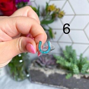 May include: A silver ring with a turquoise horseshoe design. The ring is being held by a hand.