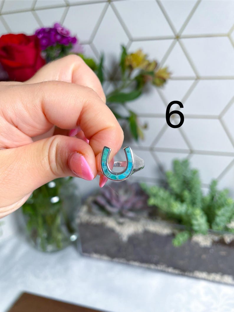 May include: A silver ring with a turquoise horseshoe design. The ring is being held by a hand.