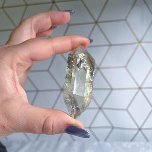 May include: A double-terminated clear quartz crystal with internal fractures and inclusions. The crystal features a hexagonal prism shape with pointed ends and displays subtle rainbow iridescence within its transparent, pale yellow structure.