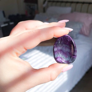 May include: A purple and white fluorite crystal palm stone. The stone is smooth and polished, with a natural, organic shape.