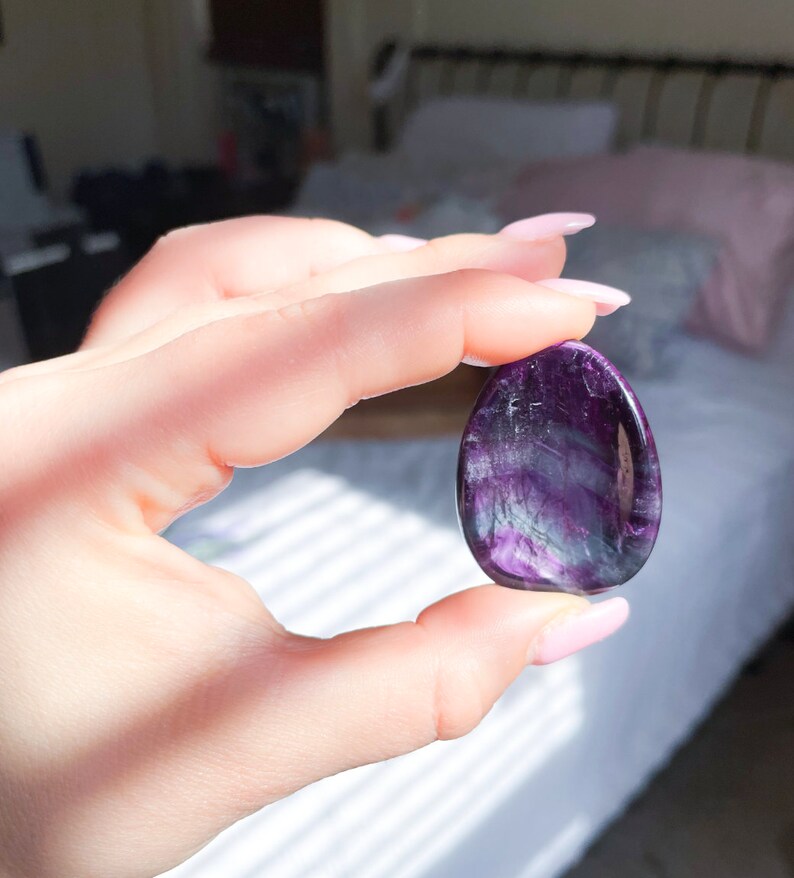 May include: A purple and white fluorite crystal palm stone. The stone is smooth and polished, with a natural, organic shape.
