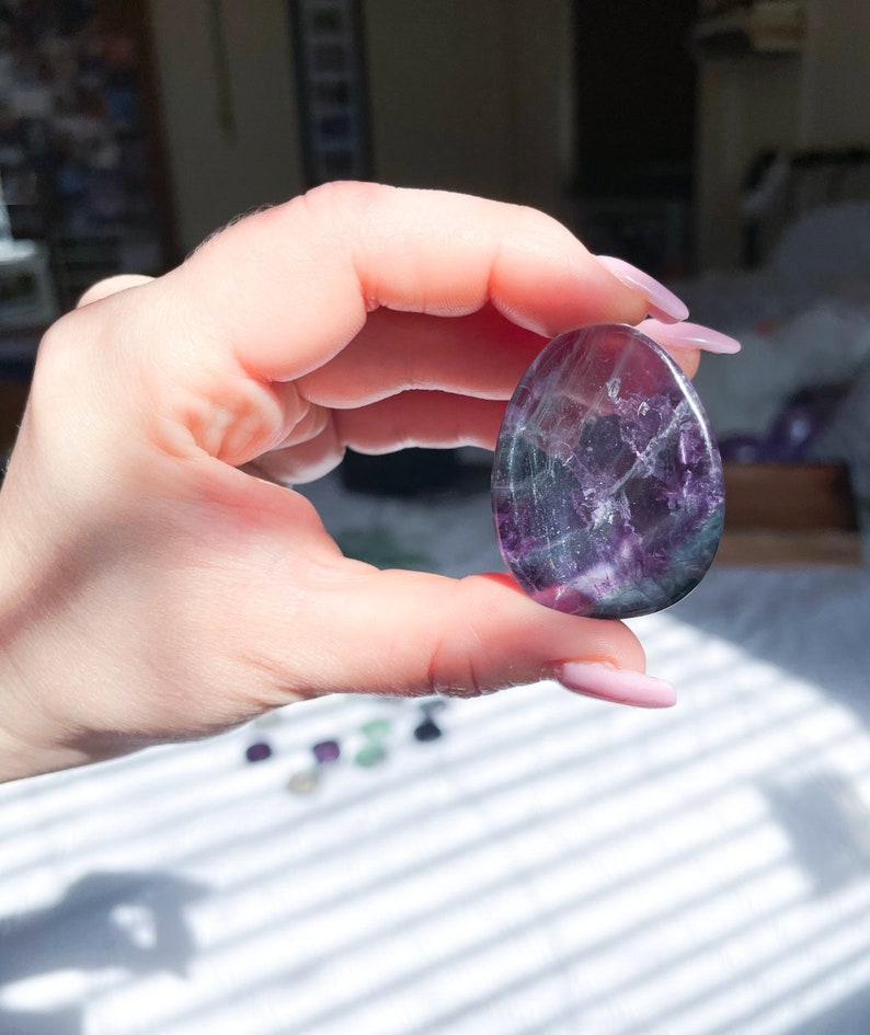 May include: A purple and green fluorite crystal egg, polished smooth. The crystal is held in a hand with manicured nails.