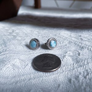 May include: A pair of silver earrings featuring a round blue gemstone set within a silver halo. The earrings are small and delicate, and the gemstone has a shimmering, iridescent effect.