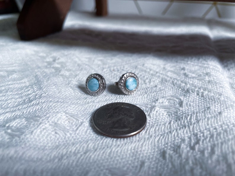 May include: A pair of silver earrings featuring a round blue gemstone set within a silver halo. The earrings are small and delicate, and the gemstone has a shimmering, iridescent effect.
