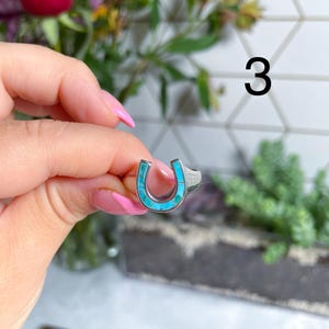May include: A silver ring with a turquoise horseshoe design. The ring is on a finger.