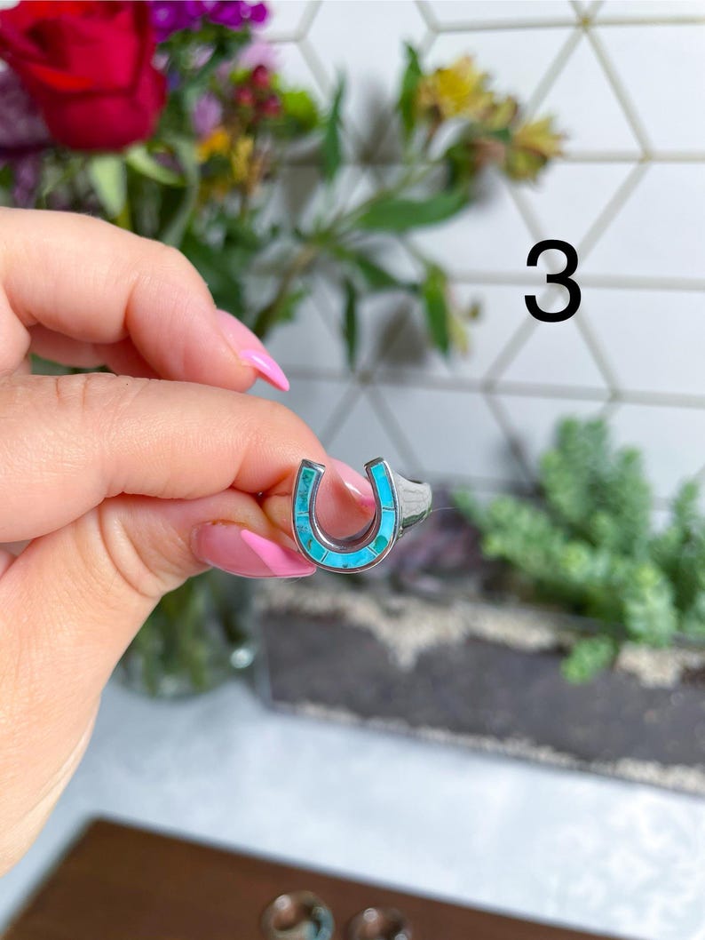 May include: A silver ring with a turquoise horseshoe design. The ring is on a finger.