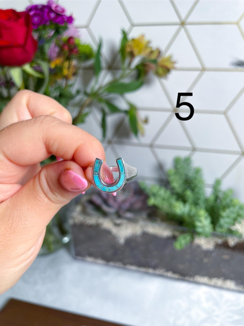 May include: A silver ring with a turquoise horseshoe design. The ring is on a finger.