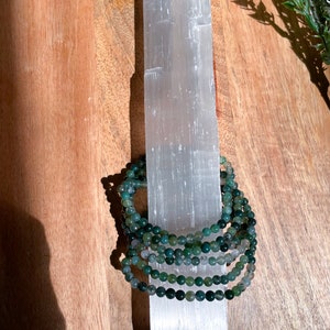 May include: A stack of green agate beaded bracelets on a white selenite charging plate. The bracelets are made of small, round beads and are stacked on top of each other.