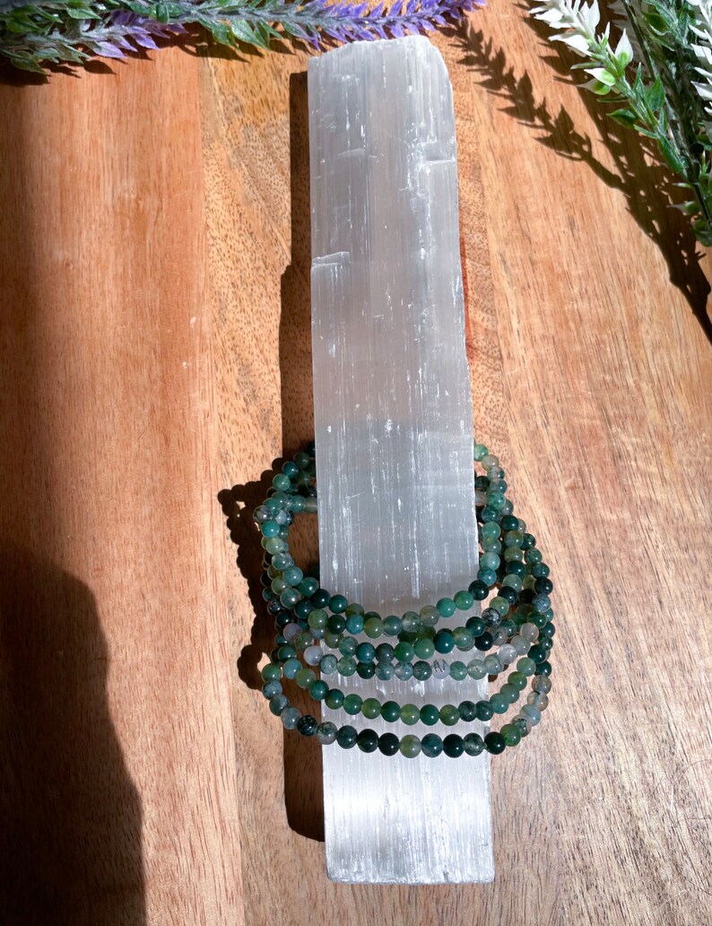 May include: A stack of green agate beaded bracelets on a white selenite charging plate. The bracelets are made of small, round beads and are stacked on top of each other.