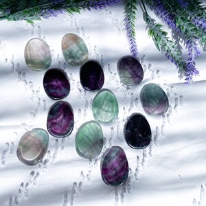 May include: A collection of 12 fluorite crystal stones in various shades of green, purple, and black. The stones are smooth and polished, and they are arranged in a random pattern on a white surface.